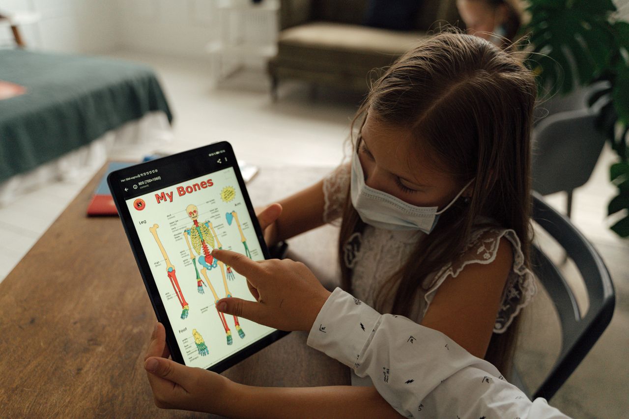 A young girl wearing a face mask uses a tablet to study anatomy at home, highlighting pandemic learning.